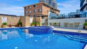 Piscina com borda azulejada azul, cercada por um edifício de tijolos e uma estrutura moderna de vidro.