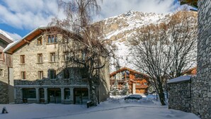 Ein verschneiter Innenhof mit Steinbauten, einem Holzauto und einem Berg im Hintergrund.