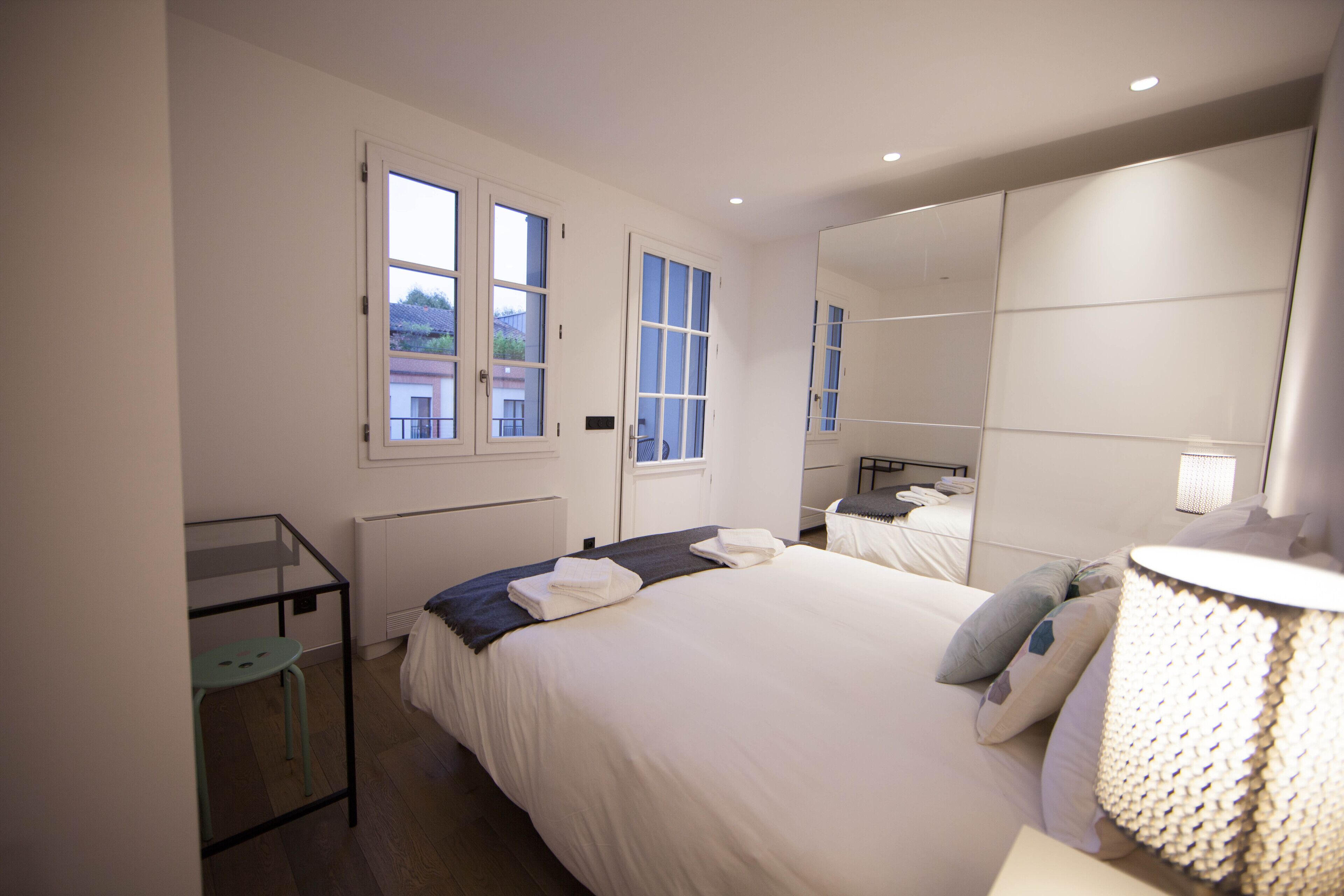 Un dormitorio con una cama grande, una mesita de noche, una silla, una ventana y un espejo.