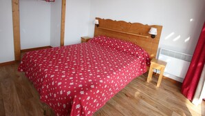 A bedroom with a wooden bed frame, a red floral bedspread, a wooden nightstand, and a wall-mounted heater. - 6-person apartment in Chamrousse, south-facing, 2 bedrooms (Chamrousse)