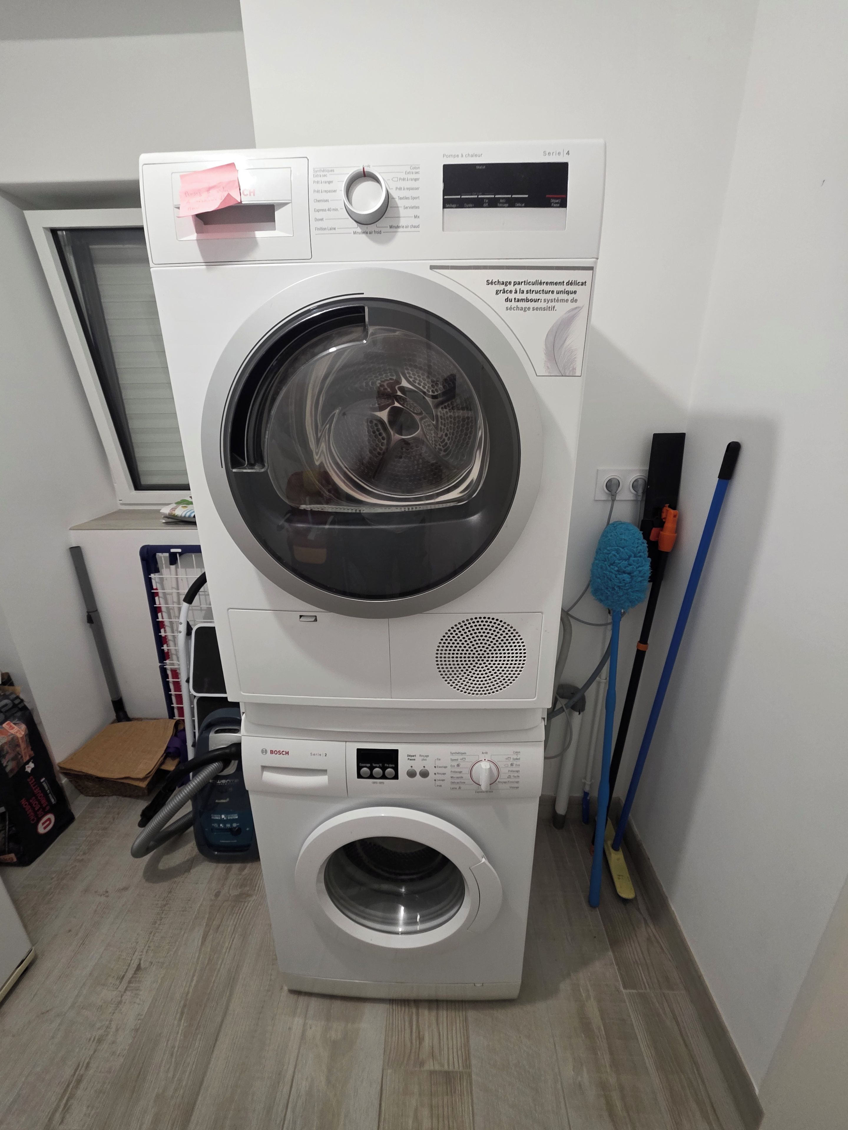 Una lavadora y secadora apiladas con un panel de control, una ventana encima de la secadora y herramientas de limpieza a un lado.