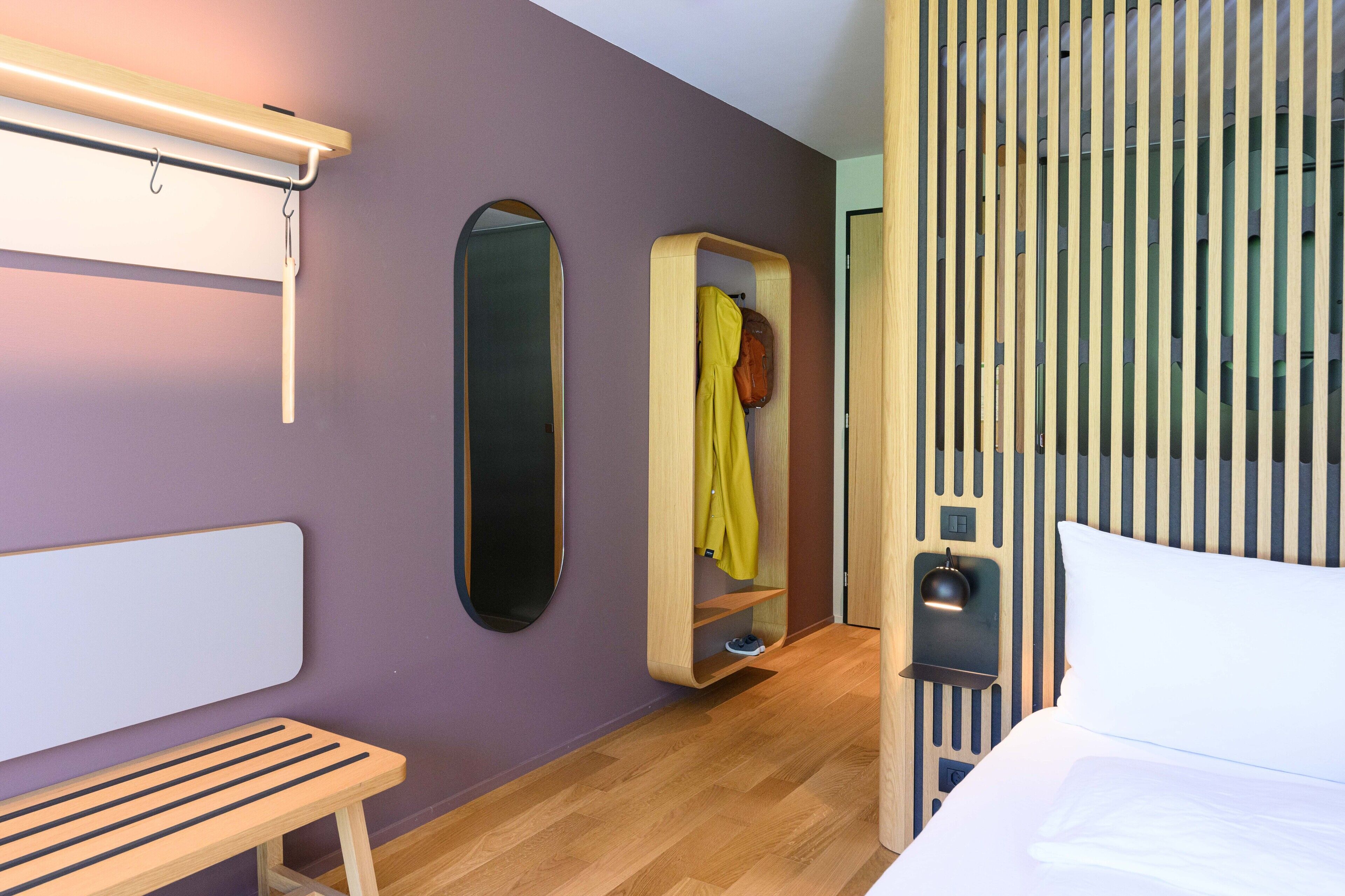 Une chambre d’hôtel moderne équipée d’un lit, d’une banquette, d’un miroir, d’une armoire et d’un luminaire fixé au mur.