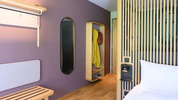 Une chambre d’hôtel moderne équipée d’un lit, d’une banquette, d’un miroir, d’une armoire et d’un luminaire fixé au mur.