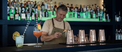 A bar with a bartender preparing drinks, a glass with a lemon slice, and a variety of bottles in the background.