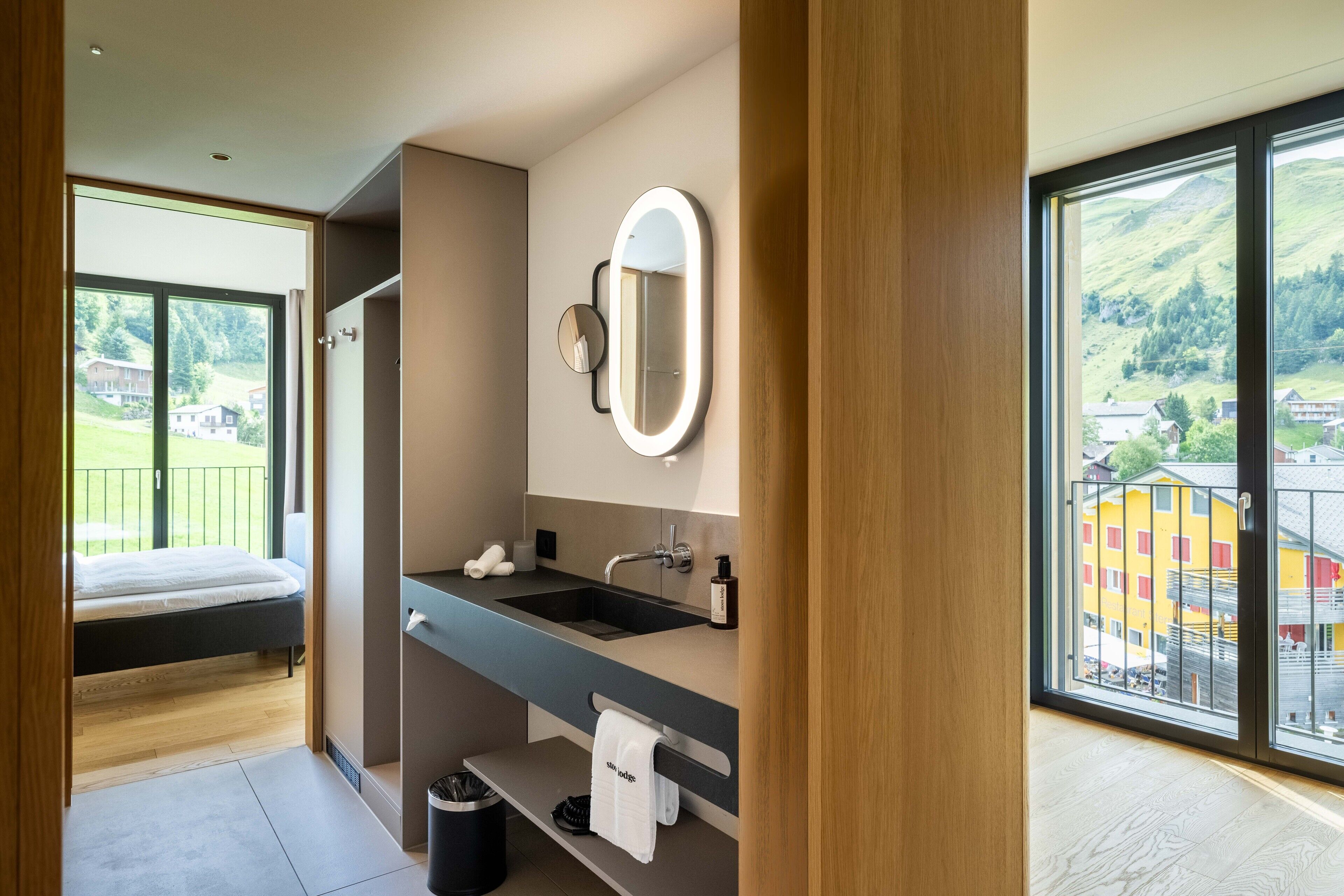 A modern bathroom with a large mirror, sink, and shower. It features a balcony with a view of a village.