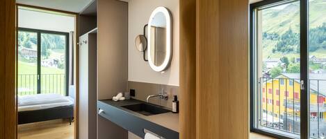 A modern bathroom with a large mirror, sink, and shower. It features a balcony with a view of a village.