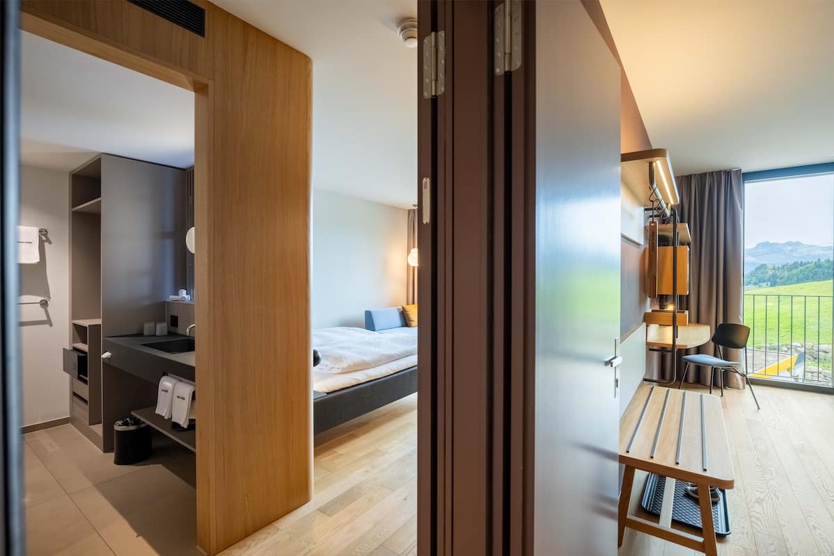 Moderni hotellihuone, jossa on sänky, yöpöytä, lamppu ja näkymä ulos.