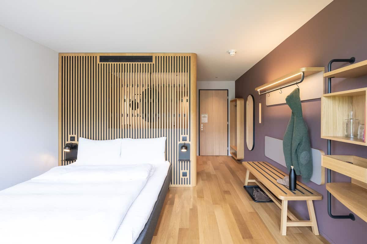 Moderni hotellihuone, jossa on suuri sänky, puupanelilla verhoiltu seinä, penkki ja hylly, jossa on hygieniatuotteita.