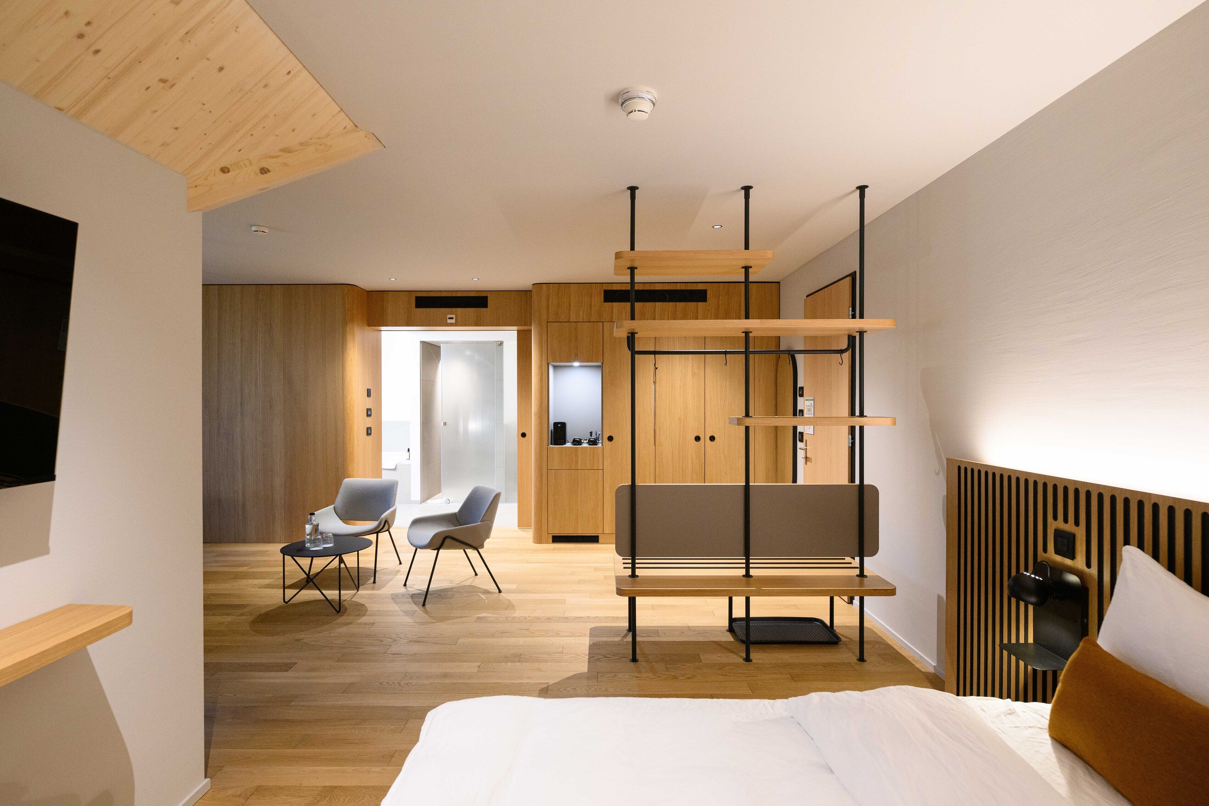 A modern hotel room with a wooden floor, a bed with white linens, a grey sofa, a small table with a chair, and a wooden cabinet.