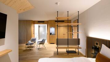 A modern hotel room with a wooden floor, a bed with white linens, a grey sofa, a small table with a chair, and a wooden cabinet.