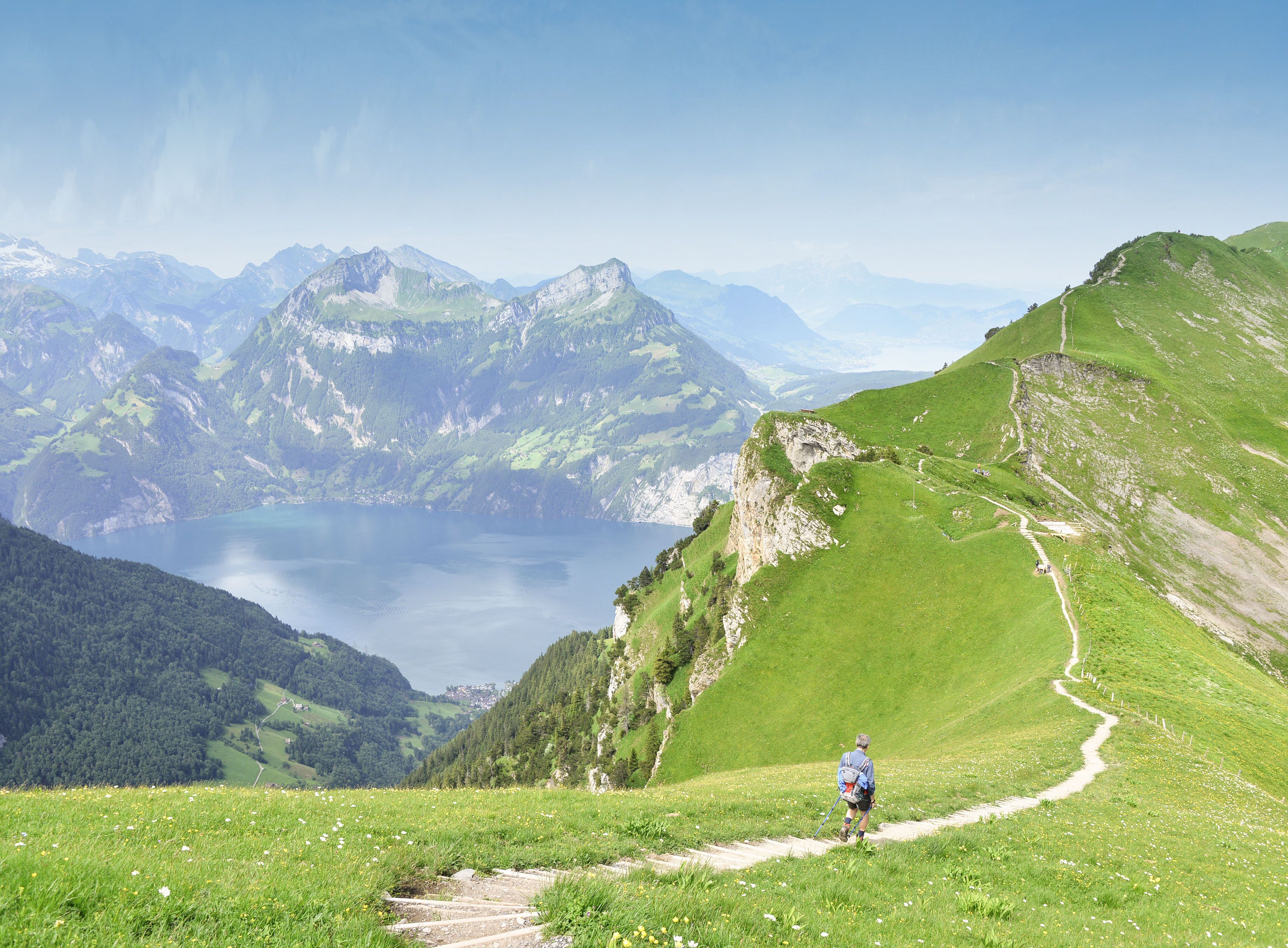 A hiker on a mountain trail with a lake and mountains in the background.