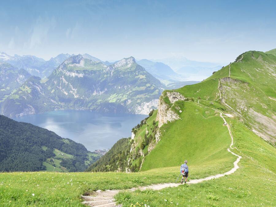 A hiker on a mountain trail with a lake and mountains in the background.