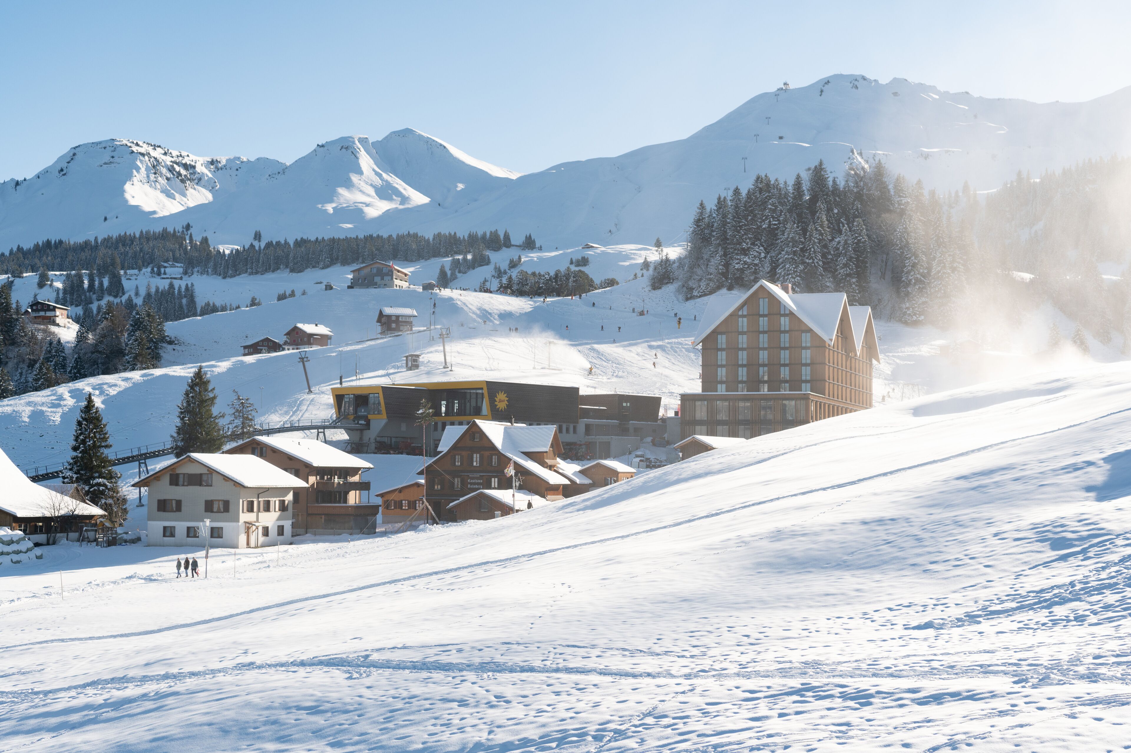 A snow-covered mountain resort with multiple buildings, including a large lodge with a prominent sign, and ski slopes in the background.