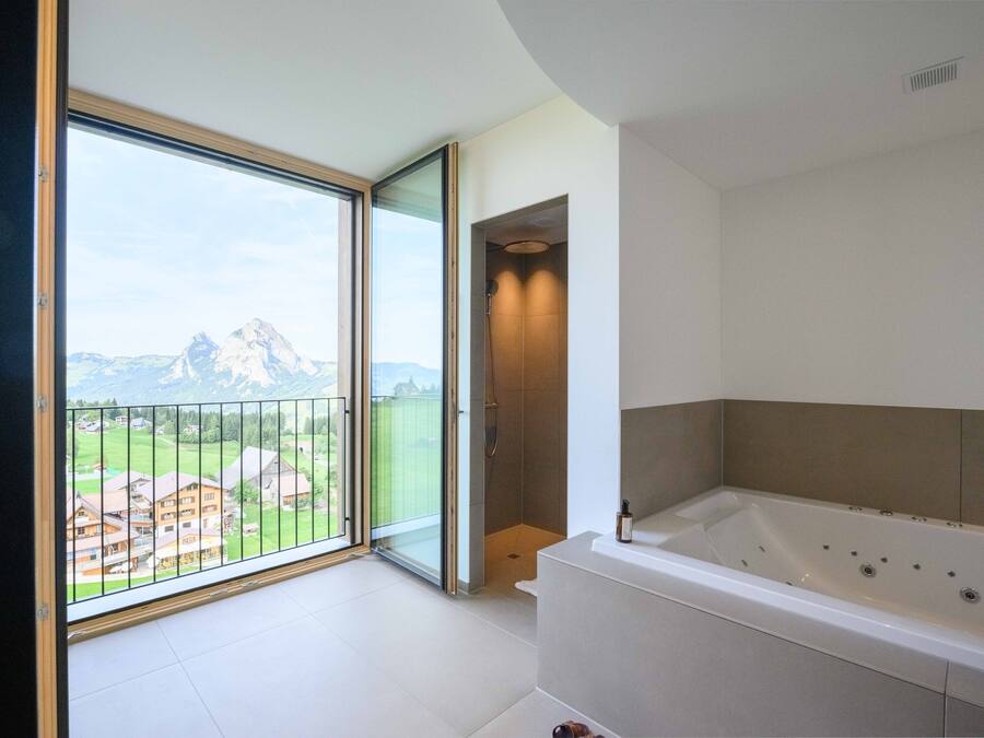 A modern bathroom with a large bathtub, a shower area, and a sliding glass door leading to a balcony with a mountain view.