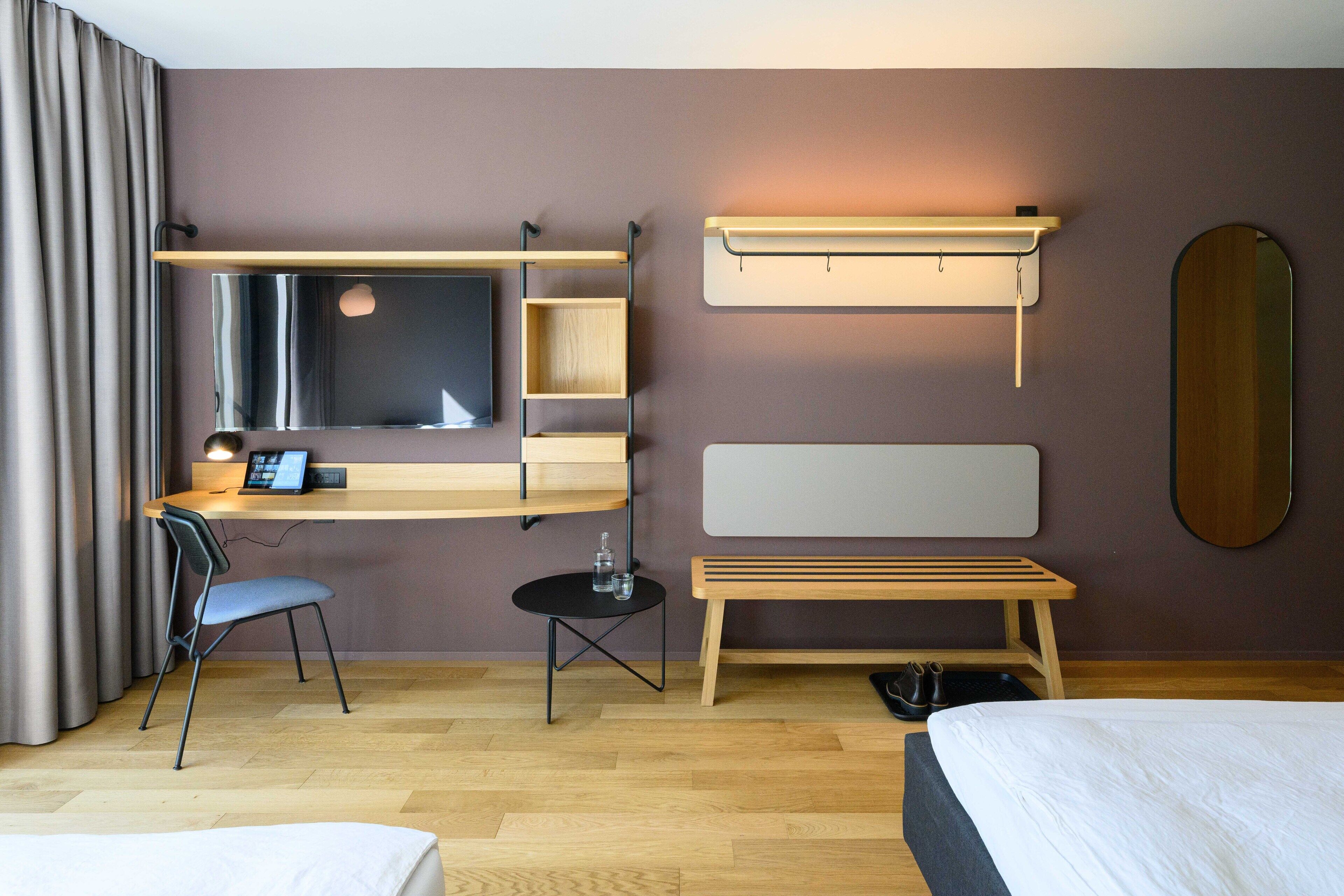 Une chambre d’hôtel comprenant un lit, un bureau avec une chaise, une petite table avec une lampe, un banc et un miroir au mur.