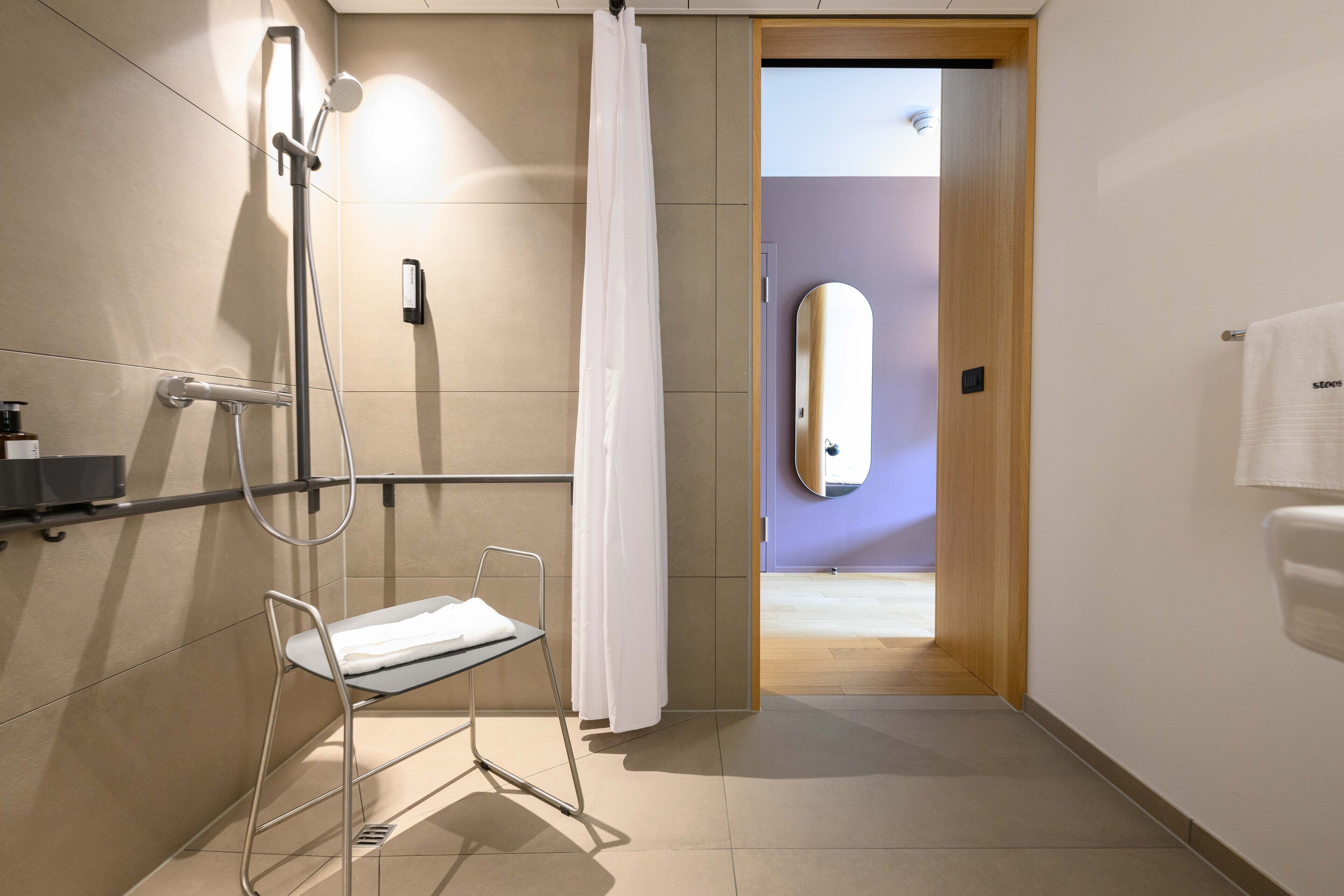 Un bany modern amb dutxa, un separador de vidre, una barra de tovalloles i una porta de fusta que dona a una altra habitació.