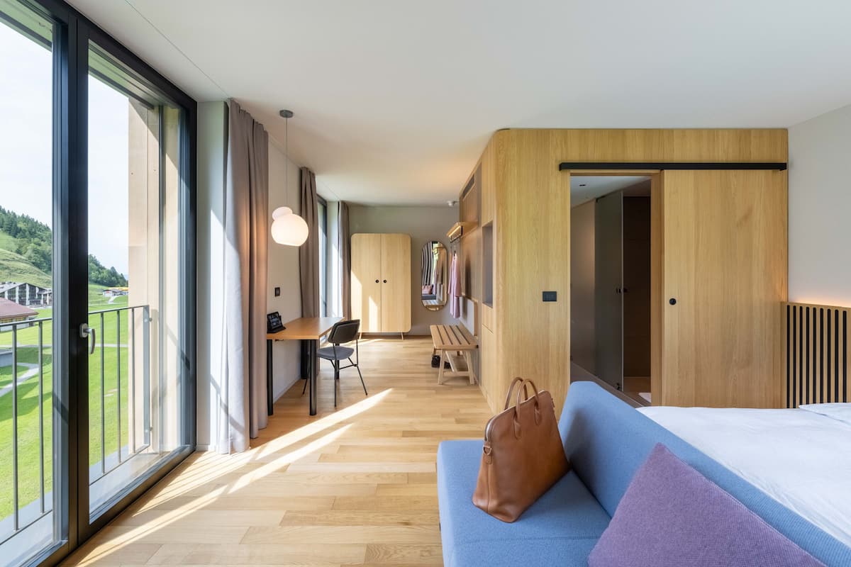 Moderni hotellihuone, jossa on suuri parveke, puulattia, sininen sohva, jonka vieressä on ruskea laukku, sekä puinen vaatekaappi.