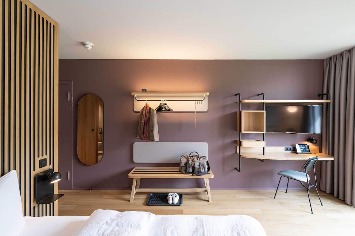 Moderni hotellihuone, jossa on sänky, penkki, televisio ja työpöytä tuolilla.
