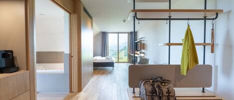 A modern hotel room with a wooden floor, a bed with a view, a desk with a laptop, and a bench with bags.