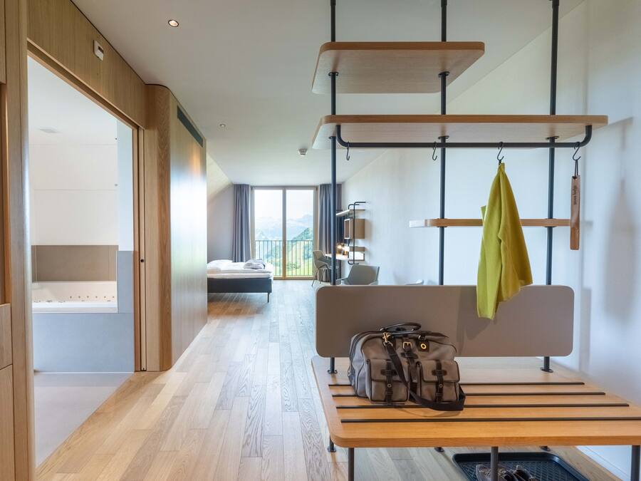 Ein modernes Hotelzimmer mit Holzboden, einem Bett mit Blick, einem Schreibtisch mit Laptop und einer Sitzbank mit Taschen.