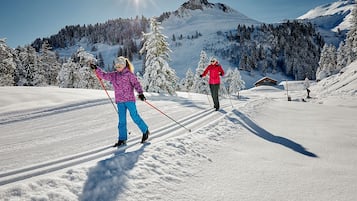 Two people cross-country skiing on a snowy path with a mountain and forest in the background.