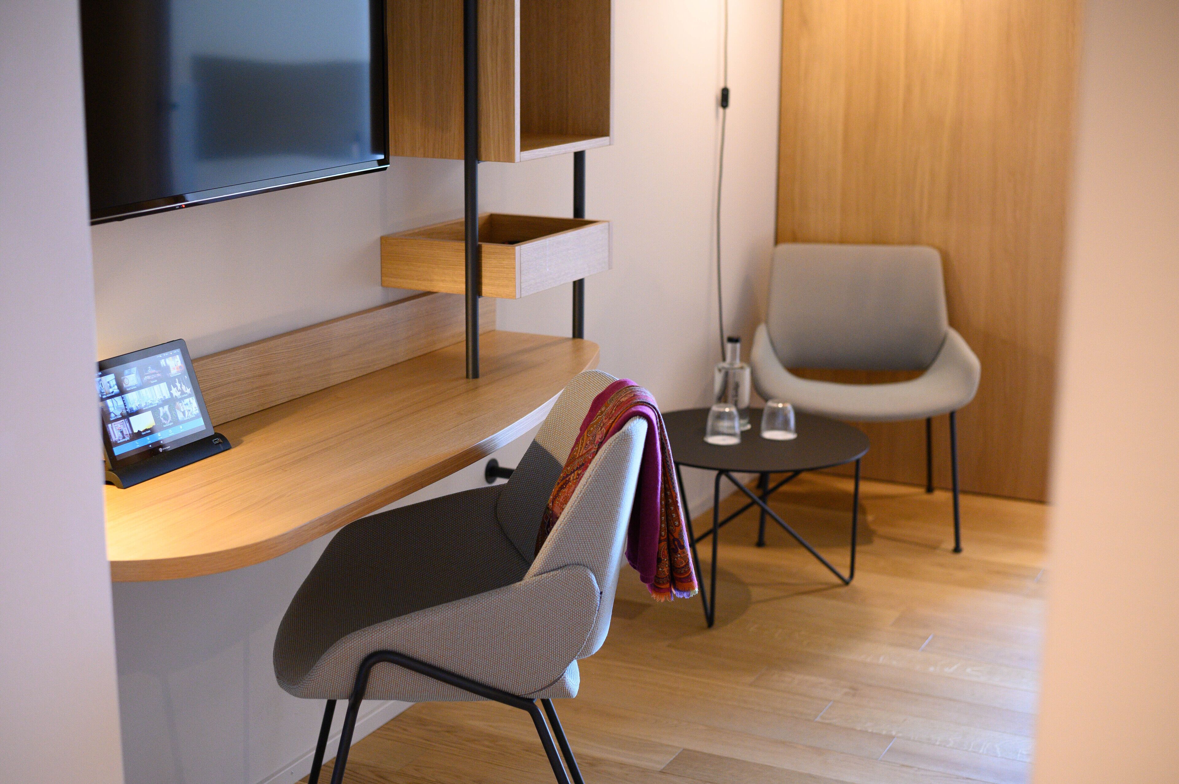 Een moderne kamer met een houten bureau, een grijze stoel, een flatscreen televisie aan de muur en een klein tafeltje met twee glaasjes.