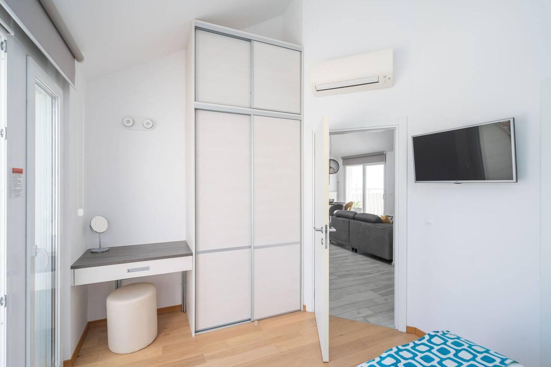 A modern, compact living space with a sliding wardrobe, a small desk, a bench, and a TV mounted on the wall.