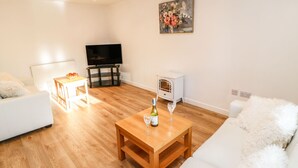 A living room with a white sofa, a wooden coffee table with a bottle and glass, a white radiator, a TV on a stand, and a small white heater. - Stable Cottage (Brampton)