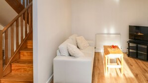 A modern living room with a white sofa, wooden staircase, and a flat-screen TV. - Stable Cottage (Brampton)