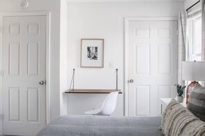 A bedroom with a bed, a chair, a bedside table, a lamp, a framed picture on the wall, and a window with curtains.