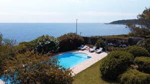 Una piscina con sillones de descanso, rodeada de vegetación y vista al mar.