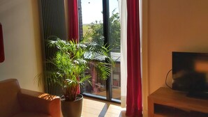 En stue med en sofa, et sofabord, et fjernsyn på et træstel, en potteplante og et stort vindue med gardiner.
