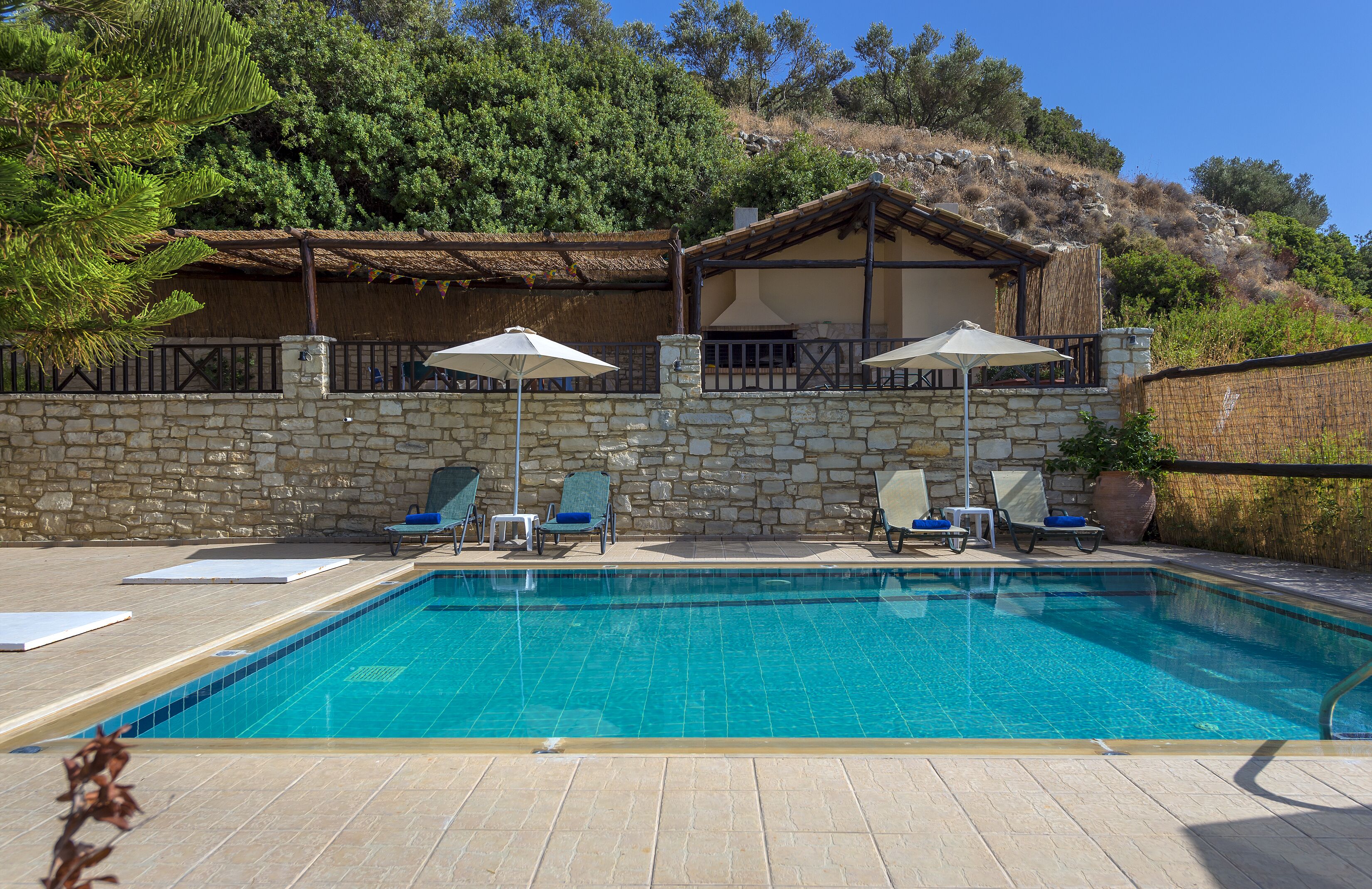 A swimming pool with lounge chairs and umbrellas, surrounded by stone walls and a hillside view.