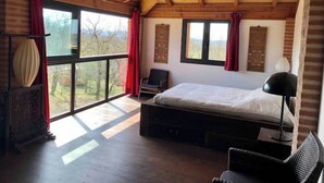 Habitación con cama, una silla, un escritorio y ventanas amplias que dan a vistas de árboles.