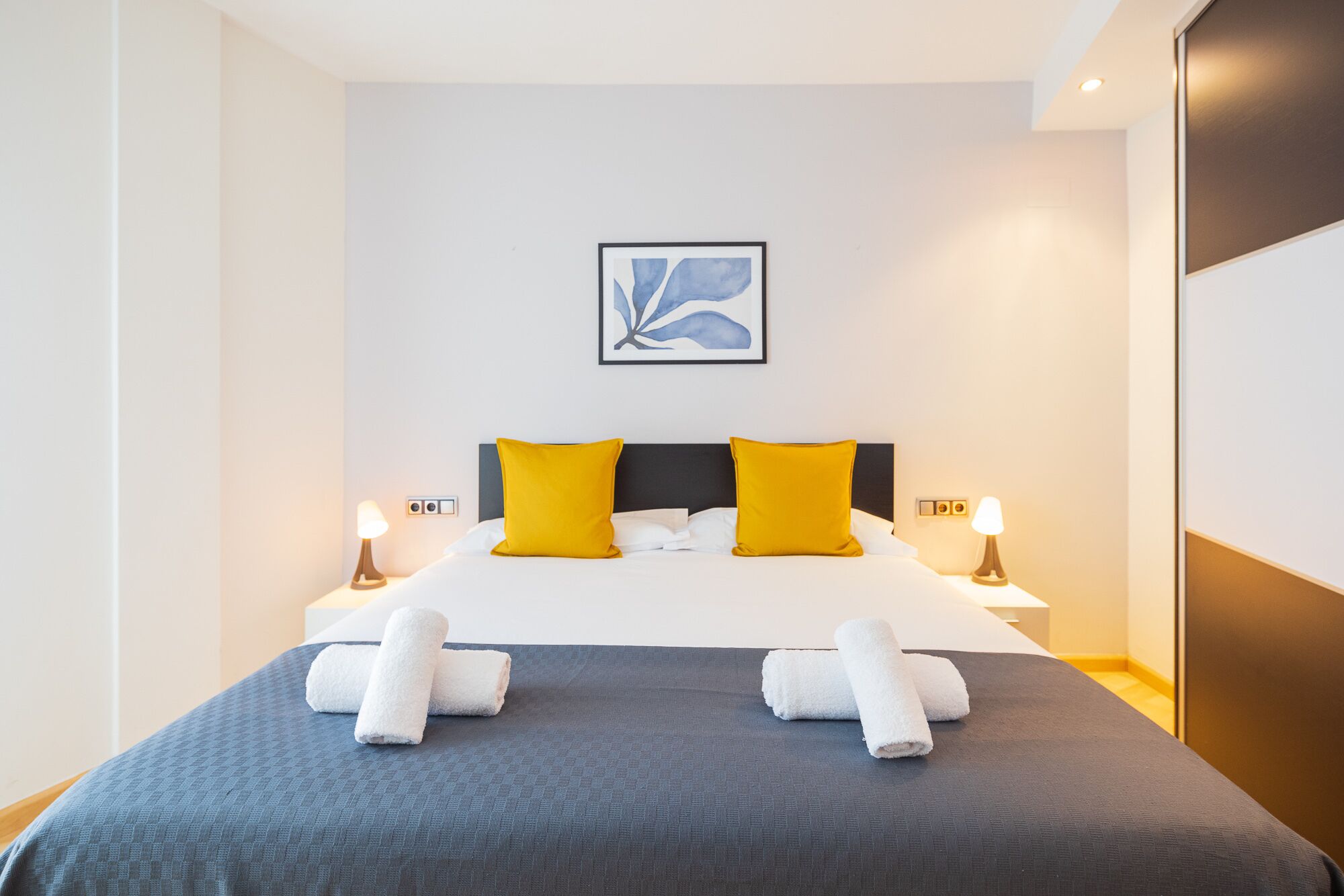 Una cama bien hecha con dos almohadas, un cuadro abstracto enmarcado encima y dos lámparas de noche.