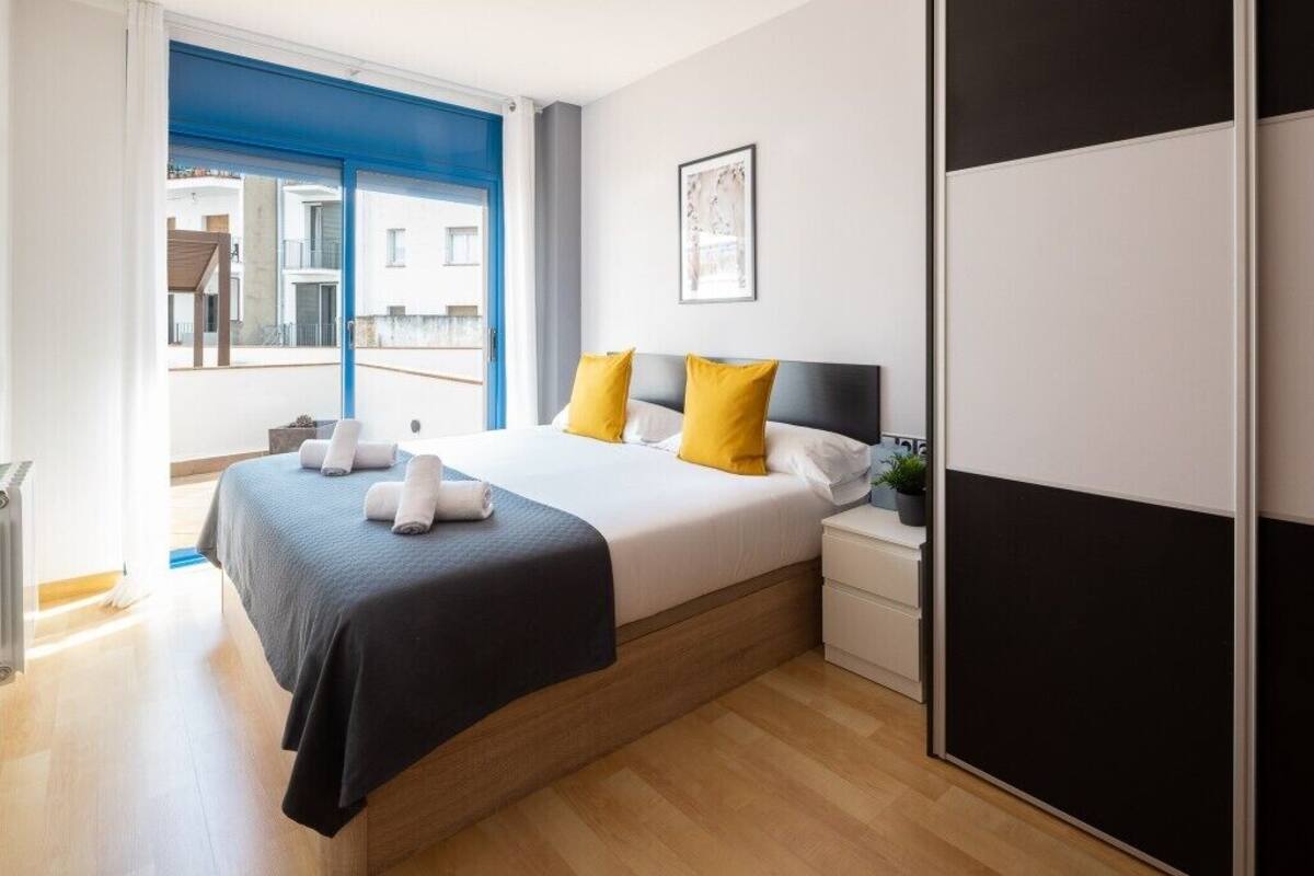 A modern bedroom with a large bed, a nightstand, a bedside table, and a view of the outside.