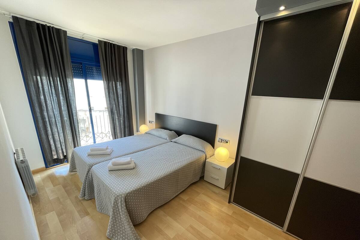 A modern hotel room with two beds, a large window with curtains, a bedside table with a lamp, and a sliding door.