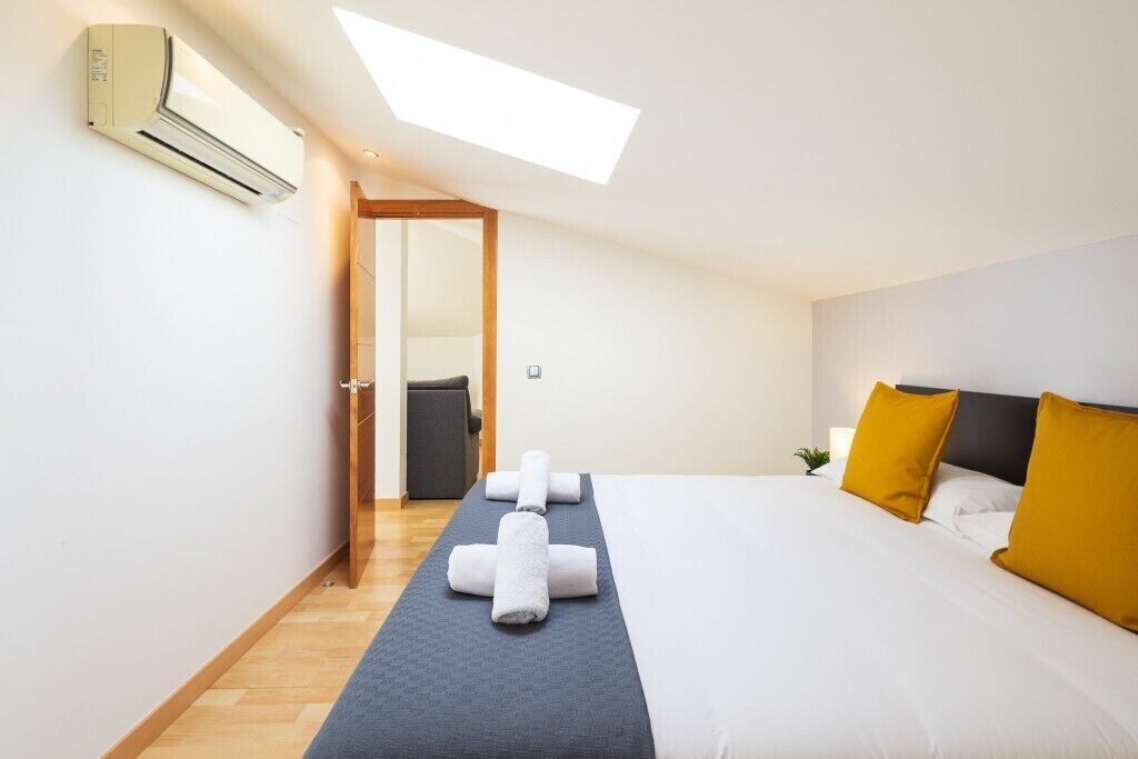 A hotel room with a skylight, a bed with white and mustard pillows, a blue towel on the floor, and an air conditioning unit on the wall.