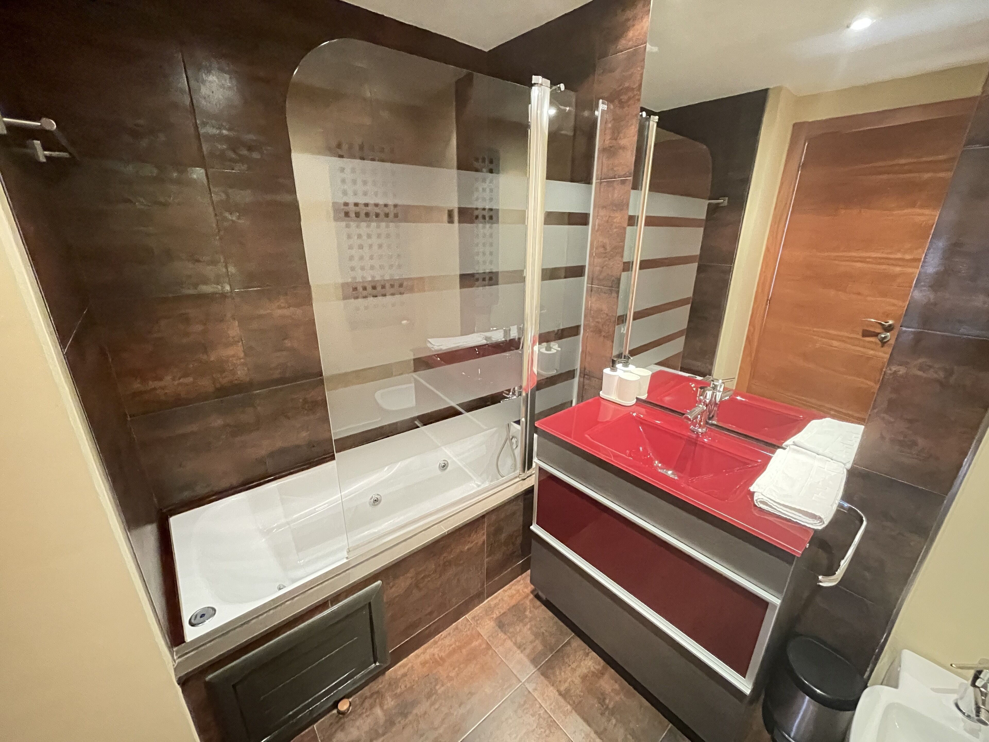 Un baño moderno con ducha envuelta en cristal, tocador con lavamanos rojo y puerta de madera.