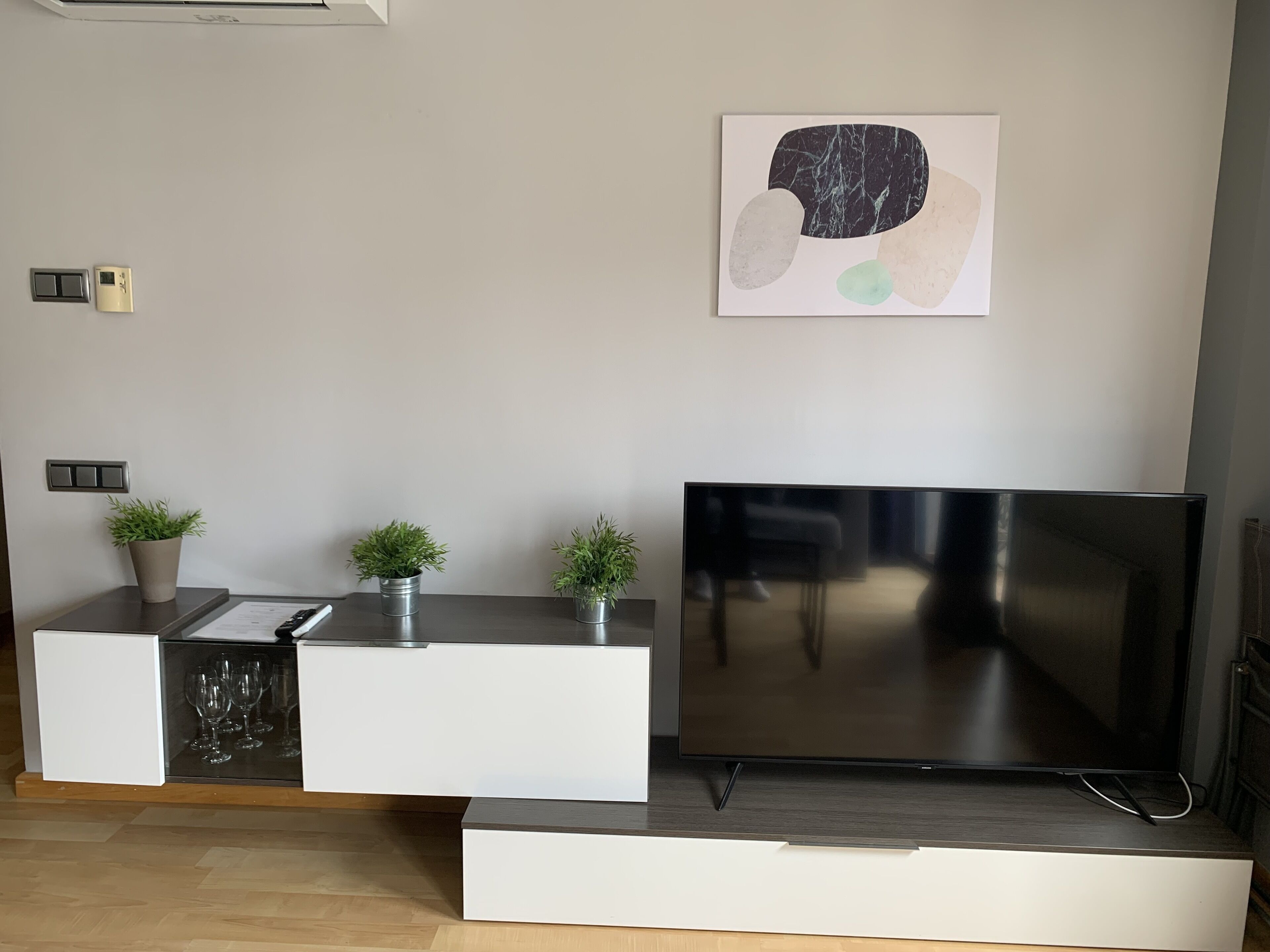 Una sala de estar moderna con un televisor de pantalla plana, un armario blanco con puertas de vidrio y dos plantas en macetas.