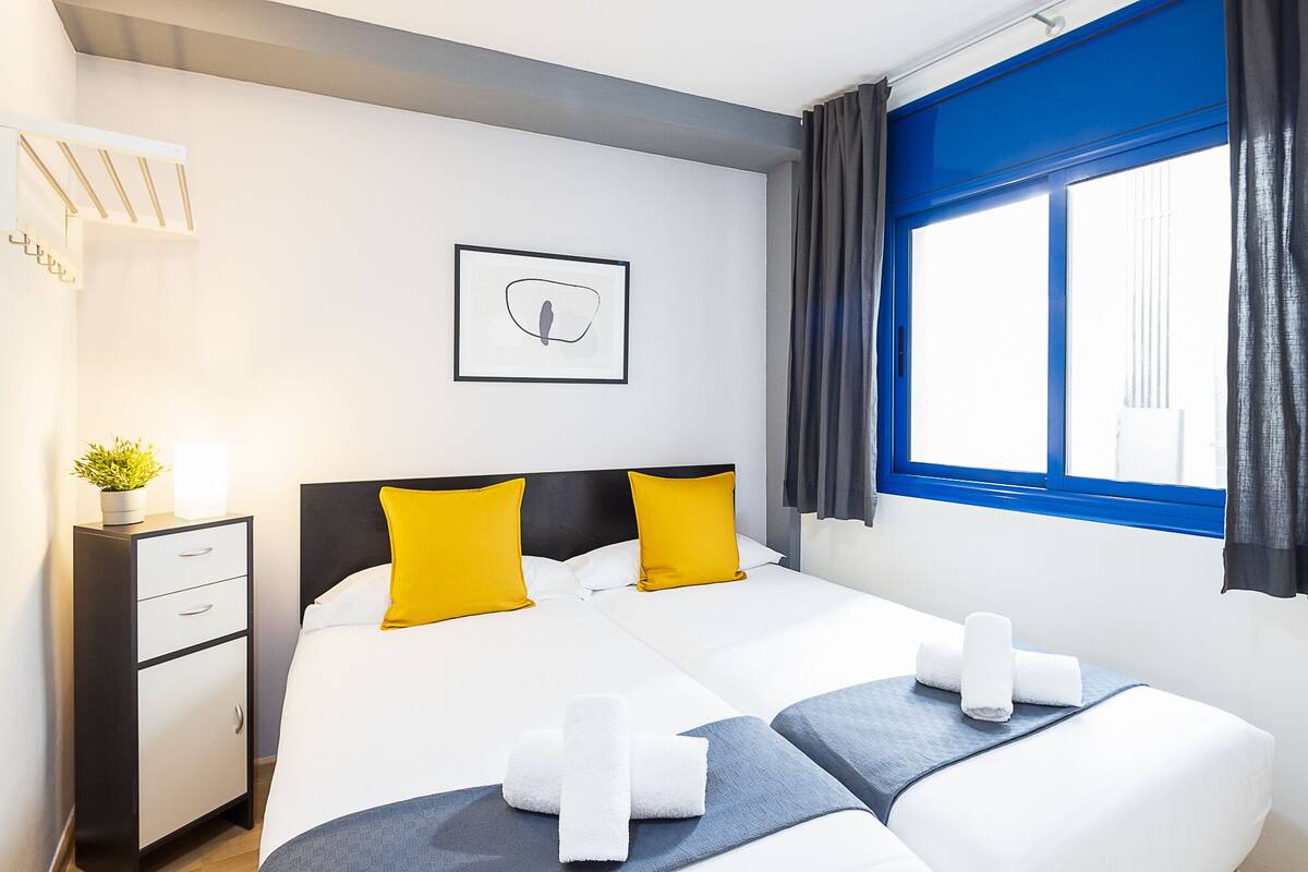 A hotel room with a bed, a nightstand with a plant, a framed picture on the wall, and a window with curtains.