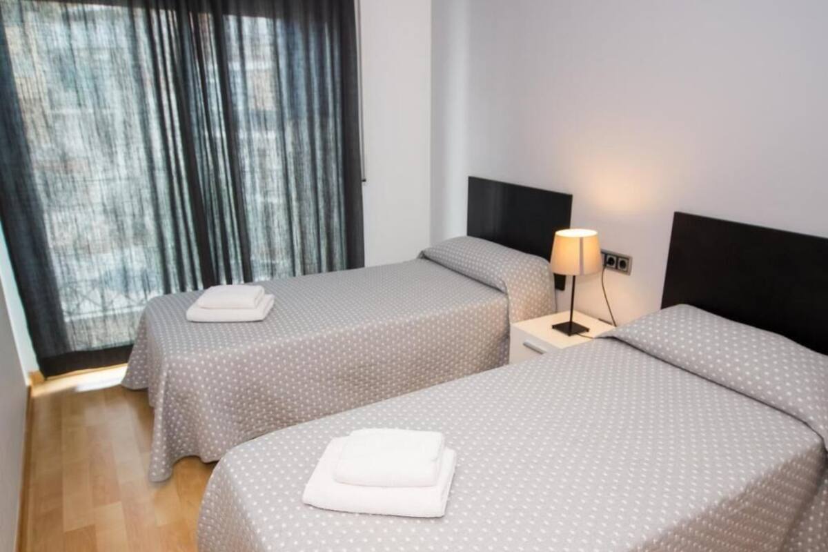A hotel room with two beds, each with a grey polka dot bedspread and a white towel folded on top. There is a nightstand with a lamp between the beds.
