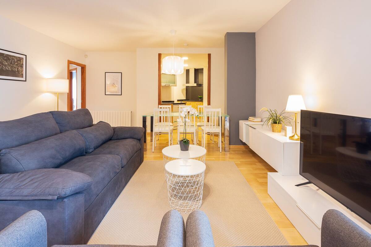 A modern living room with a grey sofa, a TV, a dining area with white chairs, and a kitchen in the background.