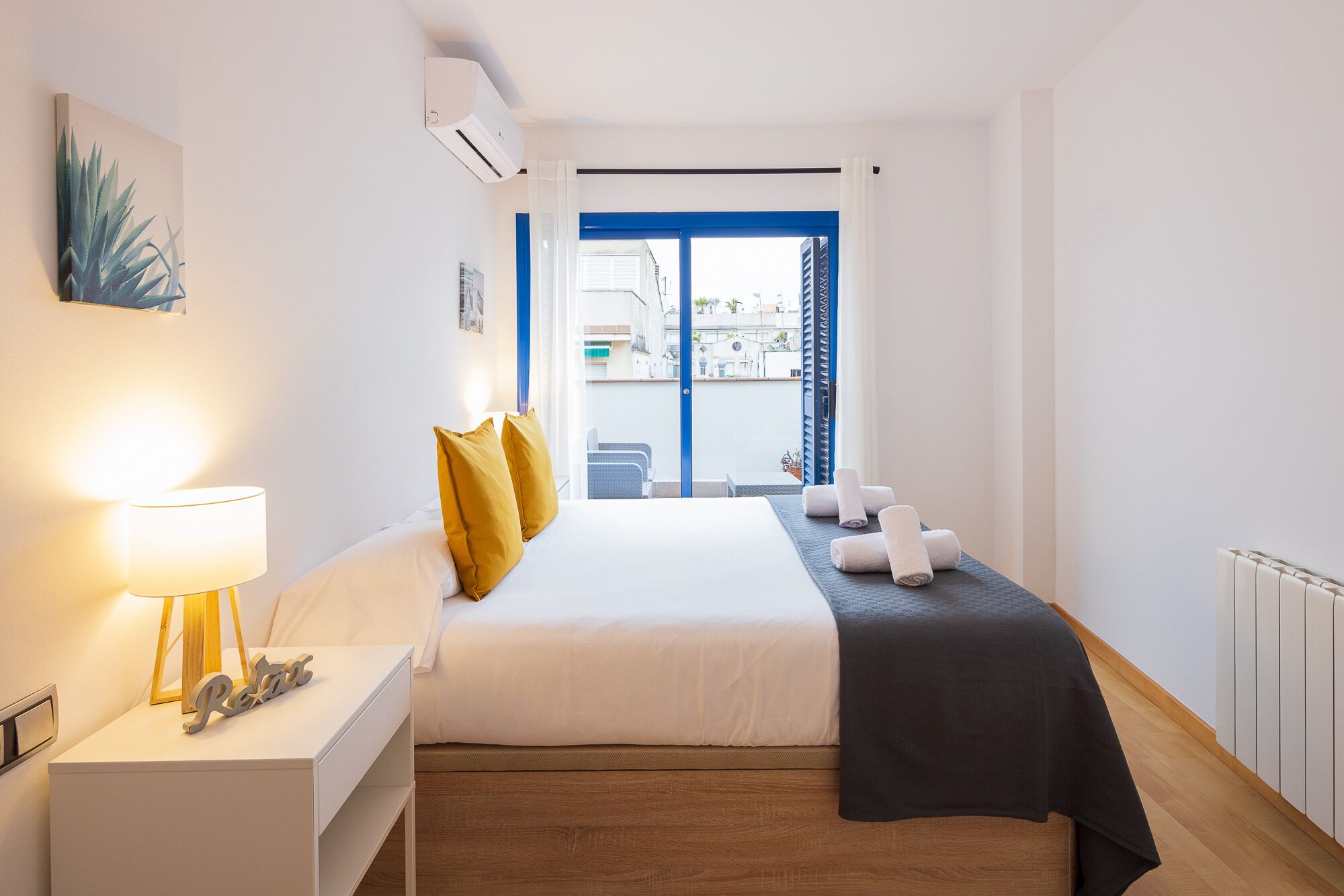 Una habitación de hotel con cama, mesita de noche con lámpara, ventana con vistas a edificios y radiador.