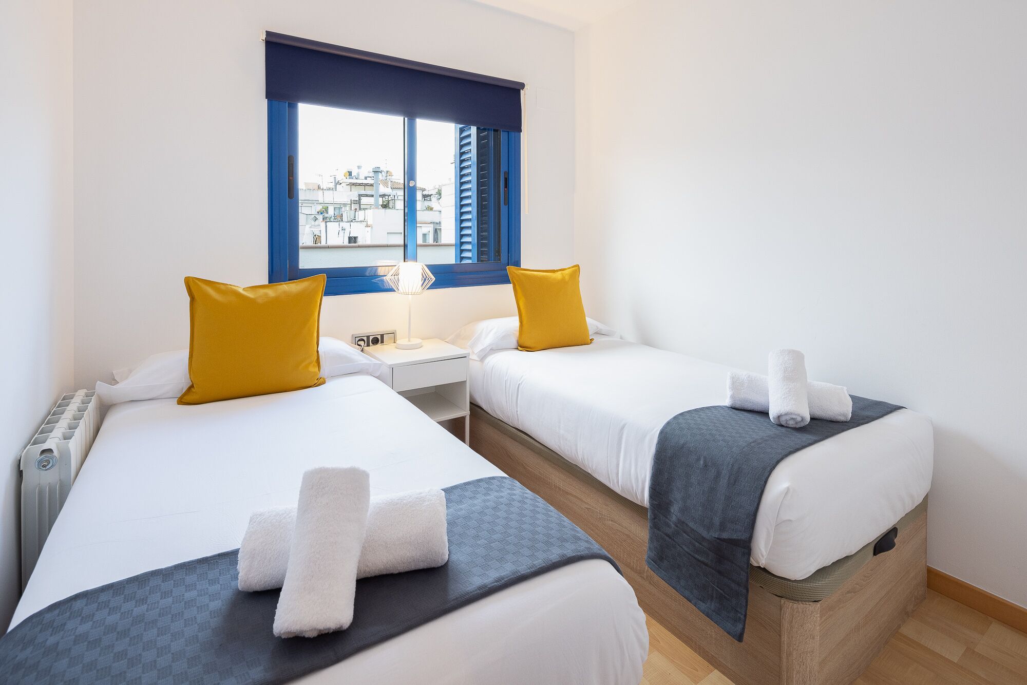 Une chambre d’hôtel avec deux lits, une fenêtre avec des volets bleus, une table de chevet avec une lampe, et une vue sur les bâtiments à l’extérieur.