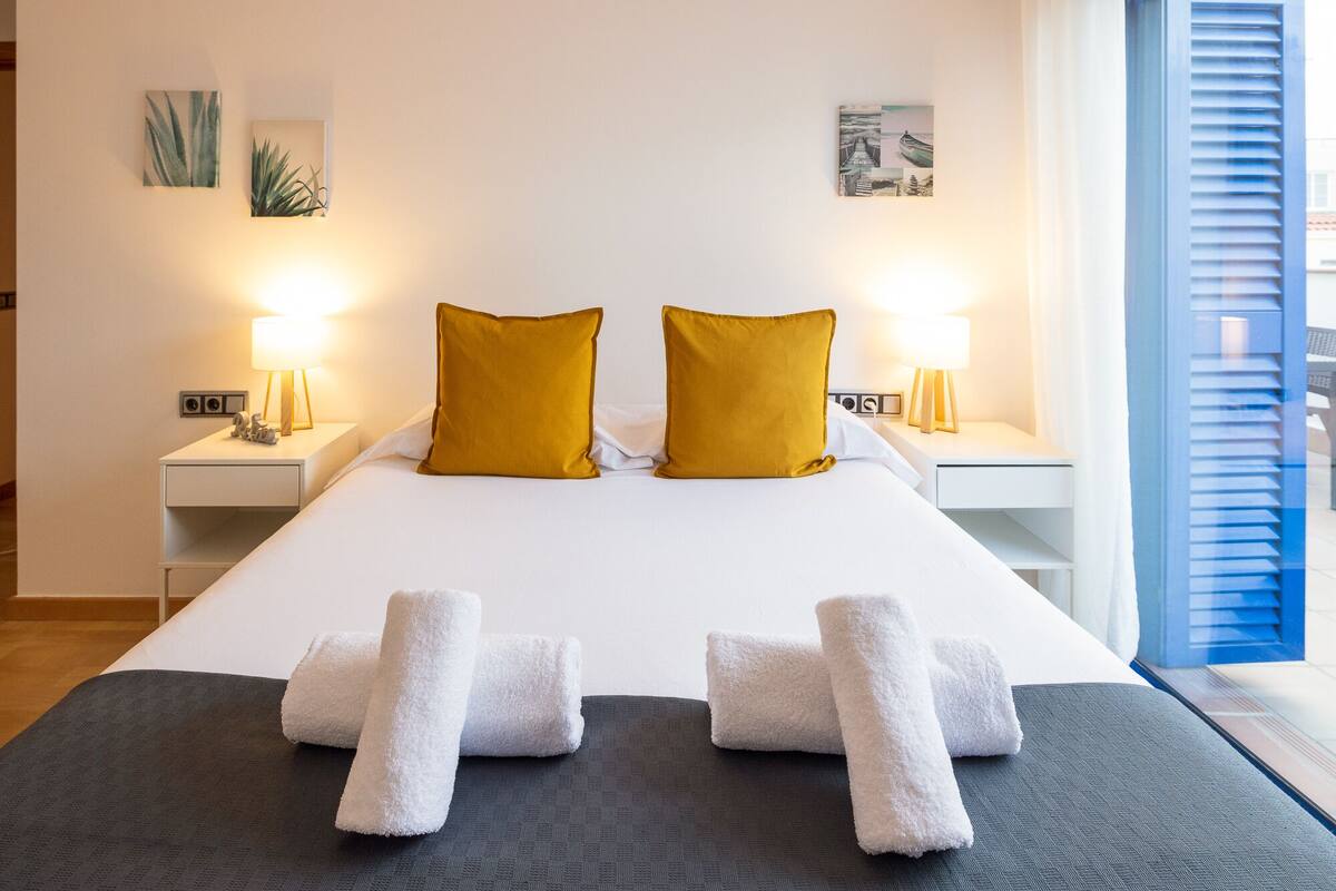 A hotel room with a large bed, two nightstands with lamps, a window with blinds, and artwork on the walls.
