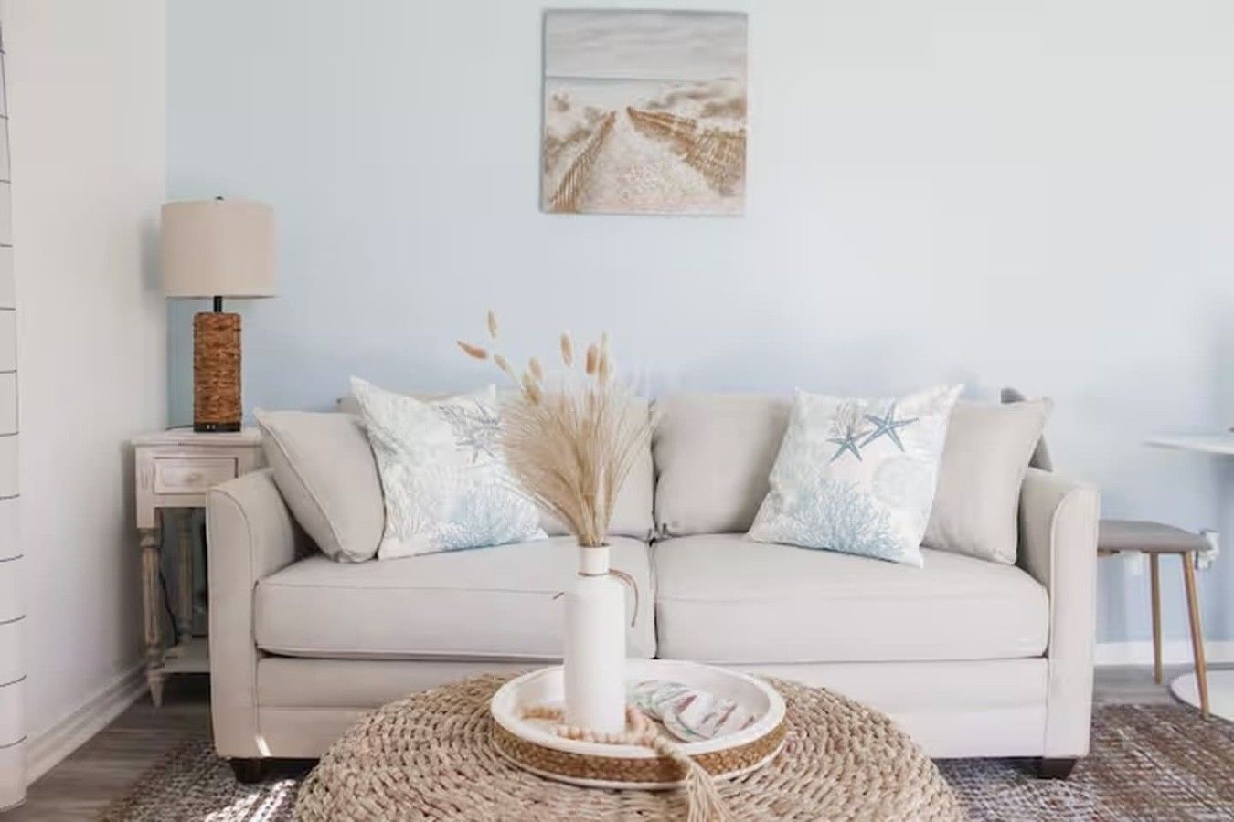 A neutral-colored sofa with decorative pillows, a woven side table, a lamp, and a wall-mounted picture.