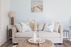 Een sofa in neutrale tinten met decoratieve kussens, een geweven bijzettafeltje, een lamp en een aan de muur bevestigd schilderij.