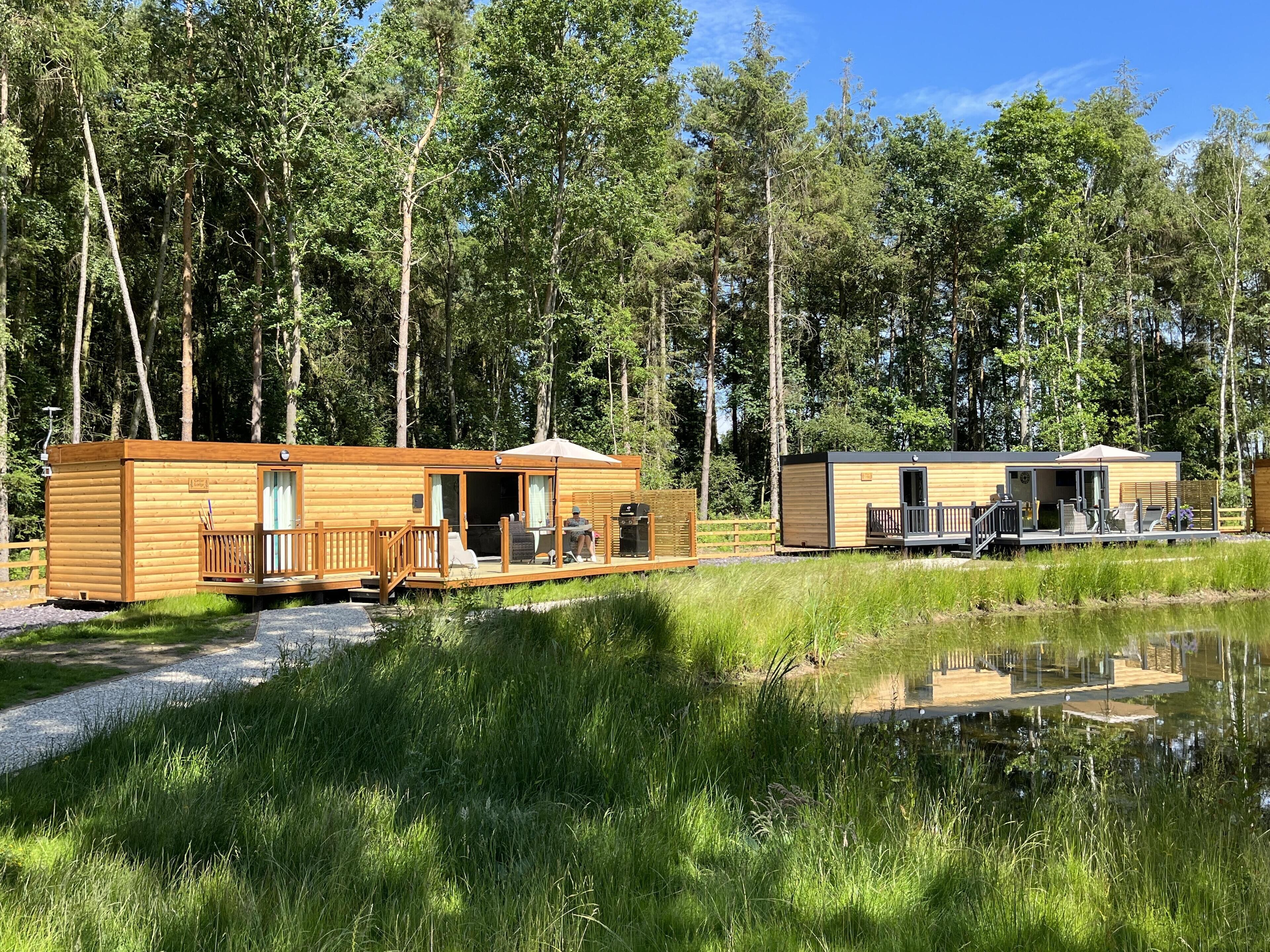 A wooden cabin with a deck and a smaller adjacent building, both surrounded by a forest and a body of water.