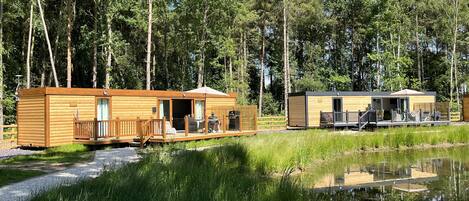 A wooden cabin with a deck and a smaller adjacent building, both surrounded by a forest and a body of water.